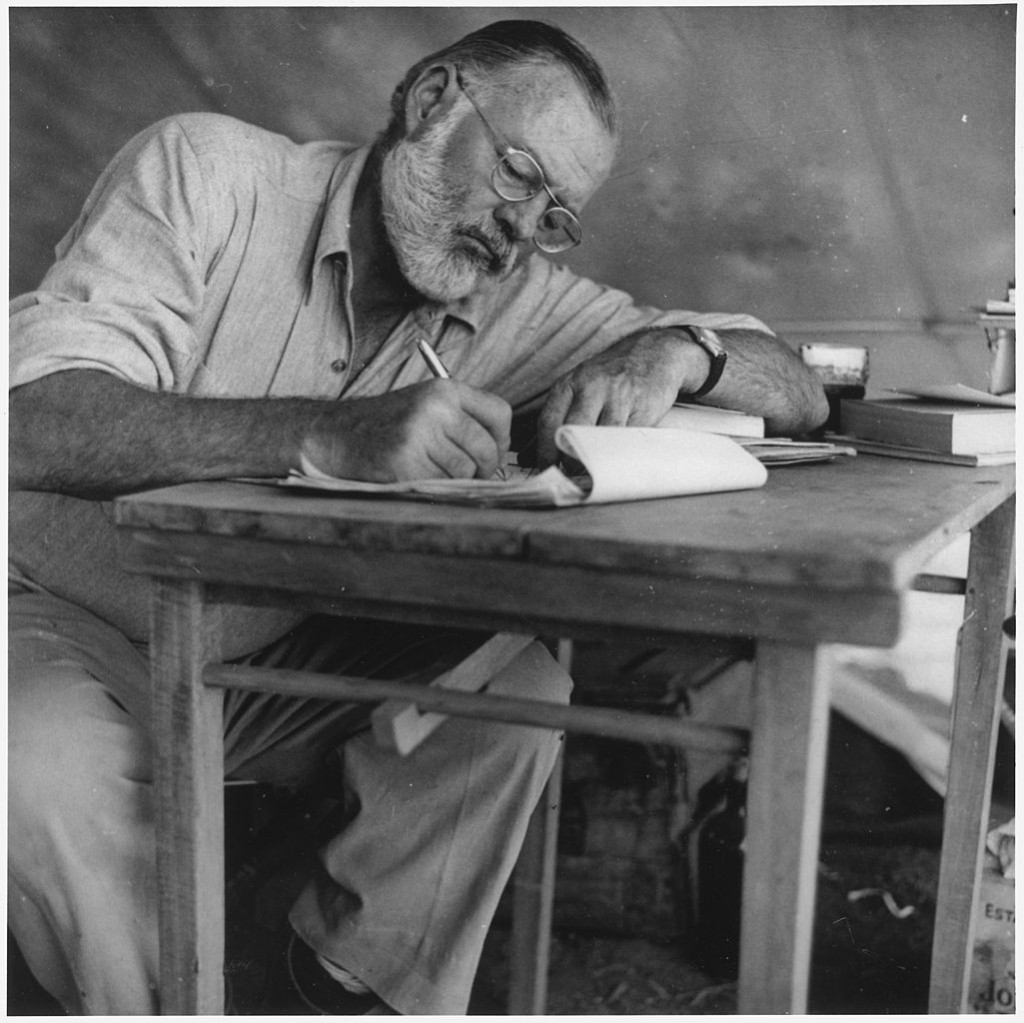 1025px-Ernest_Hemingway_Writing_at_Campsite_in_Kenya_-_NARA_-_192655
